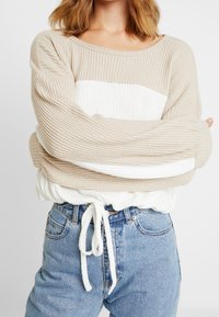 Pull en maille beige et blanc avec des manches larges, une taille élastique et un cordon de serrage. Associé à un jean taille haute bleu clair.