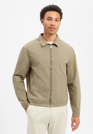 Junger Mann trägt eine beige Sweatjacke mit Reißverschluss, Kragen und elastischen Bündchen, kombiniert mit hellen Hosen, vor einem schlichten weißen Hintergrund.