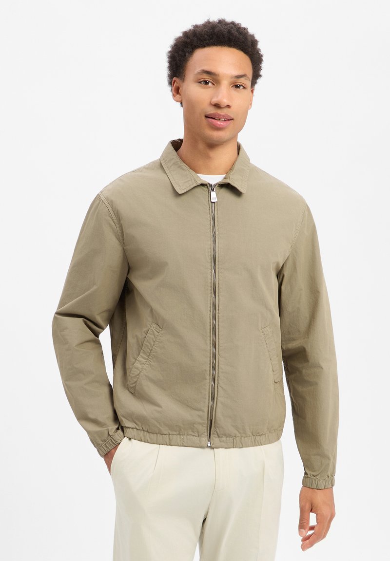 Young man wearing a beige zip-up jacket with collar and elastic cuffs, paired with light-colored pants, standing against a plain white background.