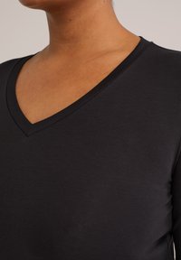 Primer plano de una persona que lleva una camiseta negra con escote en V, mostrando el cuello, la parte superior del pecho y parte del hombro contra un fondo neutral.