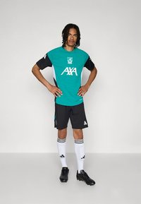 Camisola de futebol verde do Liverpool com mangas pretas, destacando detalhes em branco e logo da AXA, combinada com calções pretos e meias brancas às riscas.