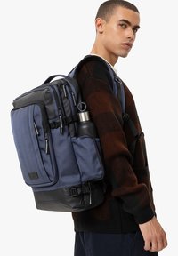 Mochila con un tejido texturizado azul y base negra. Múltiples bolsillos con cremallera, un soporte para botella de agua y tirantes ajustables.