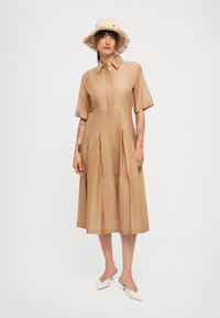 Kvinne iført en lys beige, kortermet, knelange kjole, en stråhatt, hvite spisse hæler, som står mot en enkel hvit bakgrunn.