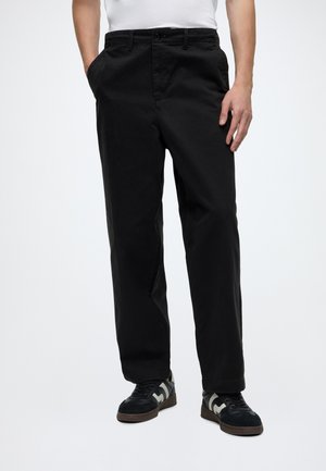 Homme portant un pantalon noir droit, des baskets noires et blanches, et une chemise blanche, debout les mains dans les poches sur fond blanc.