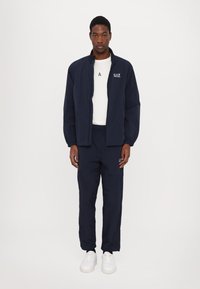 Giacca a vento blu navy con colletto rialzato, polsini elasticizzati e logo, abbinata a pantaloni blu navy con logo sulla coscia e sneakers bianche.