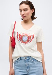 Νεαρή γυναίκα που φοράει λευκό T-shirt με πολύχρωμο σχέδιο ηλιακού φτερού, ανοιχτό μπλε τζιν, χρυσούς κρίκους σκουλαρίκια και κρατά μια κόκκινη τσάντα ώμου.