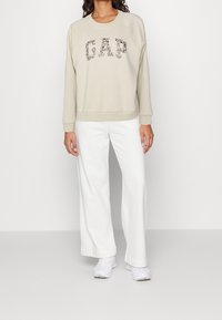 Felpa beige con "GAP" in un design floreale, scollo rotondo e maniche raglan, abbinata a pantaloni bianchi a gamba larga e scarpe bianche.
