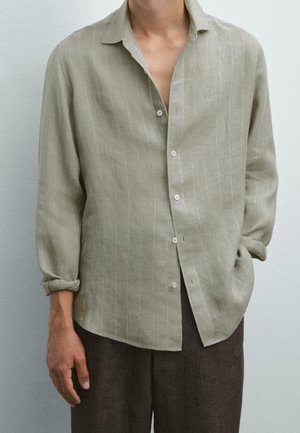 Homme portant une chemise beige clair à manches longues avec de fines rayures verticales, une main dans la poche, associée à un pantalon marron foncé.