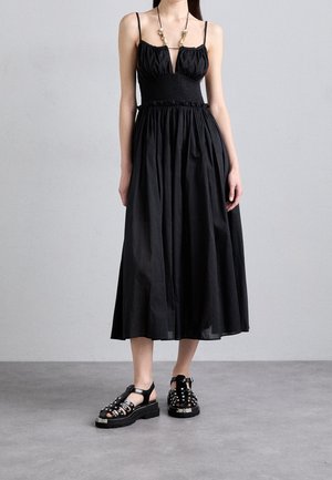 Woman wearing a black sleeveless midi dress with a smocked waist, paired with black studded platform sandals on a gray floor.