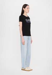 Zwarte aansluitende t-shirt met "EA7" logo in metallic tekst, gecombineerd met lichtblauwe wijde jeans en beige sneakers, staande tegen een witte achtergrond.