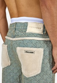 Jean hlače v svetlo modri barvi z geometrijskim vzorcem, opremljene s kremastimi umetnimi krznom obogatenimi žepi in belim pásom z črtami. Vidna oznaka "PEGADOR".