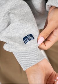 Grijze sweater met een geribbelde manchette en een marineblauw geborduurd golfontwerp op de linkermouw. Zachte stoftextuur is zichtbaar.