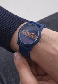 Kék szilikon karóra tiszta, kerek számlappal, nagy "GUESS" logóval fémes arany színben, arany színű órás és percmutatókkal.