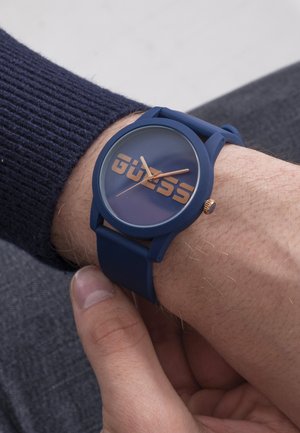 Ceas de mână din silicon albastru, cu față rotundă transparentă, logo mare „GUESS” în aur metalic și ace aurii pentru ore și minute.
