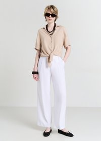 Camicetta beige a maniche corte annodata con taschino, abbinata a pantaloni bianchi a gamba larga. Occhiali da sole neri e scarpe nere texture completano il look.