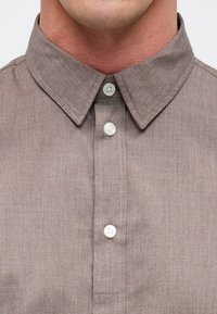 Braunes Leinenhemd mit einem Standardkragen, weißen Knöpfen und subtiler Textur, hervorgehoben durch kontrastierende Nähte entlang der Knopfleiste.