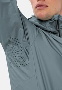 Giacca grigia chiara con zip frontale, tessuto testurizzato e un motivo a pois sulla manica sinistra per la ventilazione.