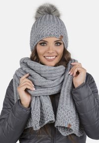 Bonnet en tricot gris clair avec un pompon et une écharpe assortie, arborant des motifs texturés et une étiquette en cuir. Les deux articles sont fabriqués en fil doux.