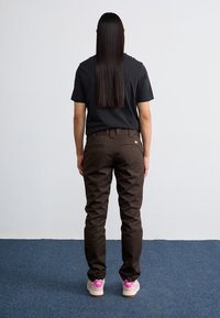 Brown cotton pants with a regular fit, featuring back pockets and a small logo. Paired with a black t-shirt and pink-accented shoes.