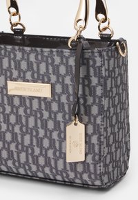 Šedá vzorovaná kabelka s charakteristickým designem loga, zlatými kovovými detaily a odnímatelnou visačkou, vyrobená z hladkého texturovaného materiálu.