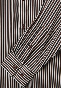 Chemise rayée avec des rayures brun foncé et crème, une texture lisse, des boutons-pression et un col pointu.