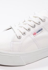 Baskets en toile blanche avec une tige texturée, un bout arrondi et des œillets argentés. Présente un logo de marque en rouge et bleu sur le côté.