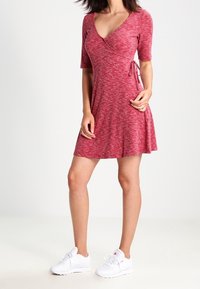 Robe portefeuille rouge à manches courtes, fabriquée dans un matériau texturé, avec un détail noué à la taille, portée avec des baskets blanches.