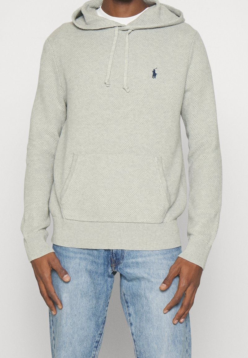 Homme portant un hoodie en maille gris clair avec une poche avant et un petit logo bleu marine, associé à un jean bleu clair, se tenant devant un fond uni.