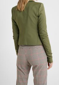 Chaqueta entallada de color verde oliva con un diseño estructurado, combinada con pantalones de cuadros de talle alto con acentos en rojo y negro.