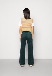 Stickad väst i orange och gula ränder över vit t-shirt, matchad med gröna kordflorsbyxor med vida ben och ljusa skor.