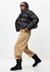 Crna puhasta jakna s krojenim dizajnom, bež široke hlače i crne čizme do koljena s kopčama. Model nosi velike sunčane naočale.