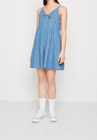 Vestido de mezclilla azul claro con tirantes spaghetti, escote en forma de V y un detalle de lazo en el pecho. Falda en capas con un flujo casual. Zapatillas blancas puestas.
