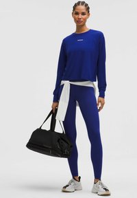 Top sportiv cu mânecă lungă de culoare albastră, cu logo, asociat cu colanți asortați. Cămașă albă legată la talie și geantă de voiaj neagră. Pantofi sport.