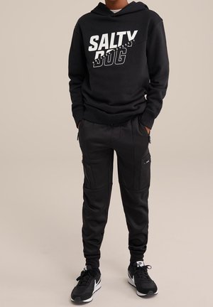 Sweat à capuche noir avec "SALTY DOG" imprimé en blanc, associé à un pantalon de jogging noir avec des poches zippées, alliant tissu et texture.