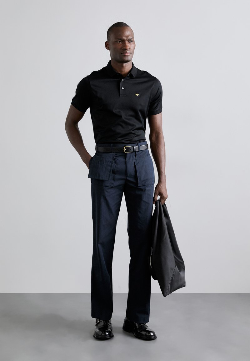 Schwarzes Poloshirt mit kurzen Ärmeln und dezentem Logo; kombiniert mit navyfarbigen Cargo-Hosen mit Taschen. Das Modell hält eine schwarze Tasche.