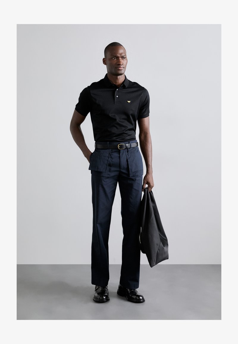Black polo shirt with short sleeves and a subtle logo; paired with navy cargo pants featuring pockets. The model holds a black bag.