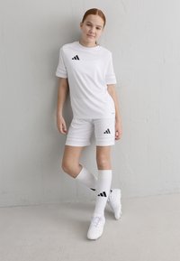 Weißes Sportoutfit, bestehend aus einem kurzärmeligen Shirt, Shorts und knielangen Socken, mit schwarzen Adidas-Logo-Akzenten und strukturiertem Stoff.