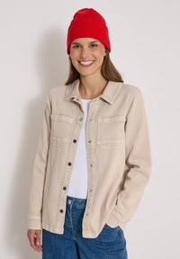 Femme souriante portant un bonnet rouge, une veste beige boutonnée sur une chemise blanche et un jean bleu, debout devant un fond clair uni.