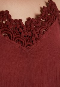 Top burdeos con un diseño de escote en V, que presenta un intrincado borde de encaje en el cuello y una tela texturizada, mostrando una ligera apariencia arrugada.
