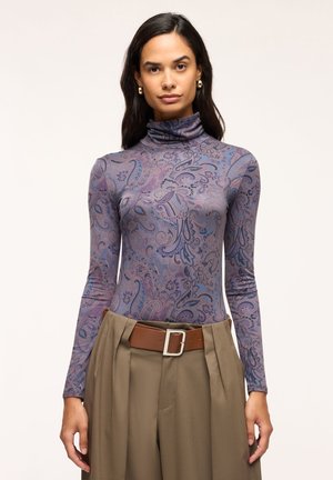 Donna che indossa un dolcevita aderente a maniche lunghe viola con fantasia paisley e pantaloni beige a vita alta con cintura marrone, in piedi su uno sfondo semplice.