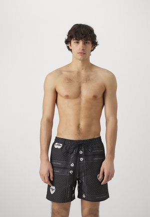 Bañador negro con un patrón de puntos, acentos en blanco, cintura elástica con cordón y detalles en parches. El material parece ligero.
