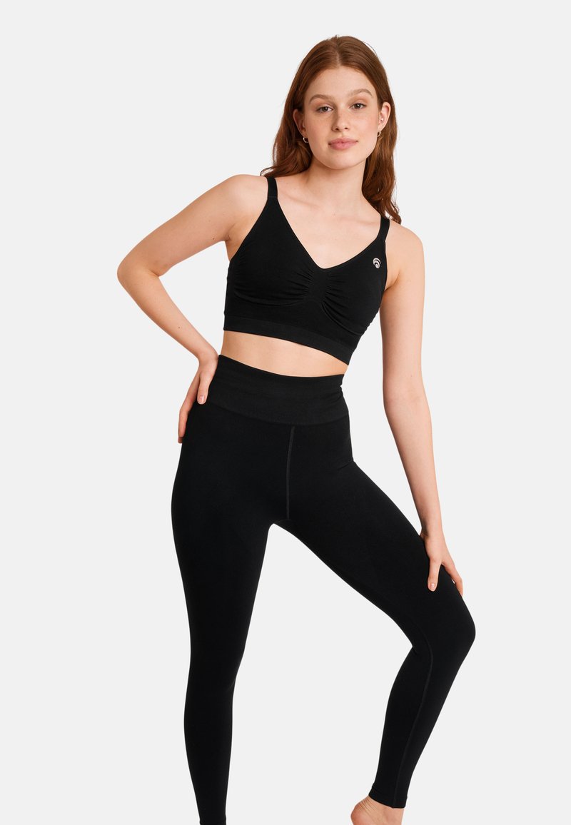 Czarny stanik sportowy z dekoltem w kształcie litery V i marszczonym detalem, zestawiony z czarnymi legginsami o wysokim stanie. Oba wykonane z gładkiego, elastycznego materiału.
