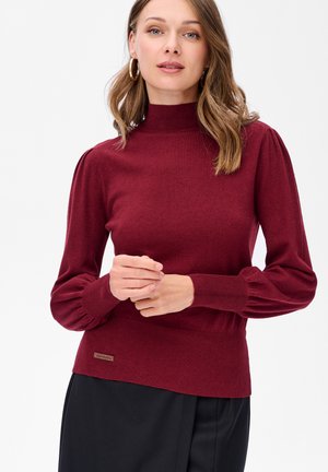 Kobieta w bordowym swetrze o wysokim kołnierzu z bufkami na długich rękawach oraz złotymi kolczykami w kształcie kół, stojąca na tle białym.