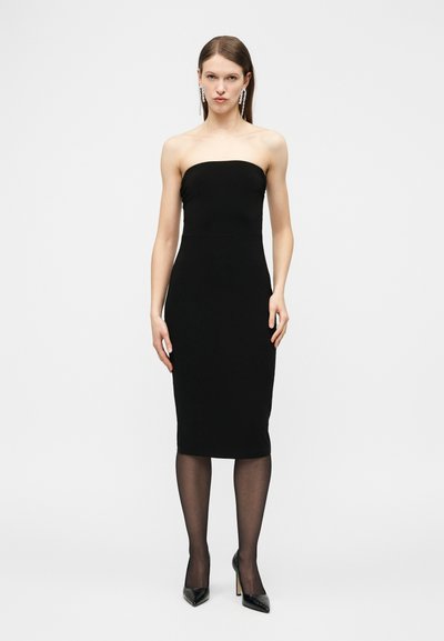 Γυναίκα με μαύρο midi φόρεμα χωρίς τιράντες, μαύρες διάφανες καλσόν, μαύρες μυτερές γόβες και μακριά κρεμαστά σκουλαρίκια, που στέκεται μπροστά σε ένα απλό φόντο.