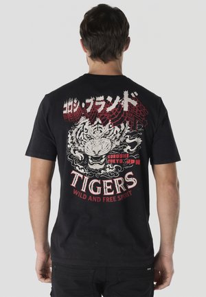 Hombre con camiseta negra mostrando diseño trasero con tigre rugiente, texto japonés y las palabras "TIGERS Wild and Free Spirit" en rojo y blanco.