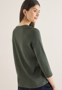 Chemise à manches longues vert foncé avec un col arrondi, tissu doux et coupe décontractée. Détails de coutures visibles le long des épaules et des manches.