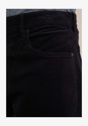 Musta corduroy-housut, joissa on teksturoitu pinta, varustettuna tavallisilla taskuilla ja hienovaraisilla metalliosilla vyötärön lähellä.