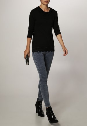 Kobieta idąca, ubrana w czarną bluzkę z długim rękawem, szare obcisłe dżinsy, czarne błyszczące botki, trzyma w prawej ręce małą czarną kopertówkę.