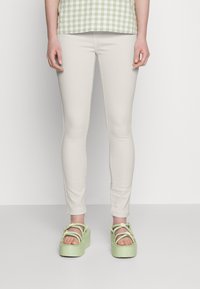 Jeans justos bege claro combinados com sandálias de plataforma verdes chunky, apresentando detalhes florais e várias tiras. Top com padrão não mostrado.