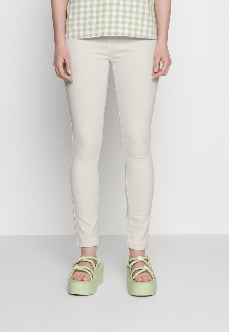 Jeans justos bege claro combinados com sandálias de plataforma verdes chunky, apresentando detalhes florais e várias tiras. Top com padrão não mostrado.